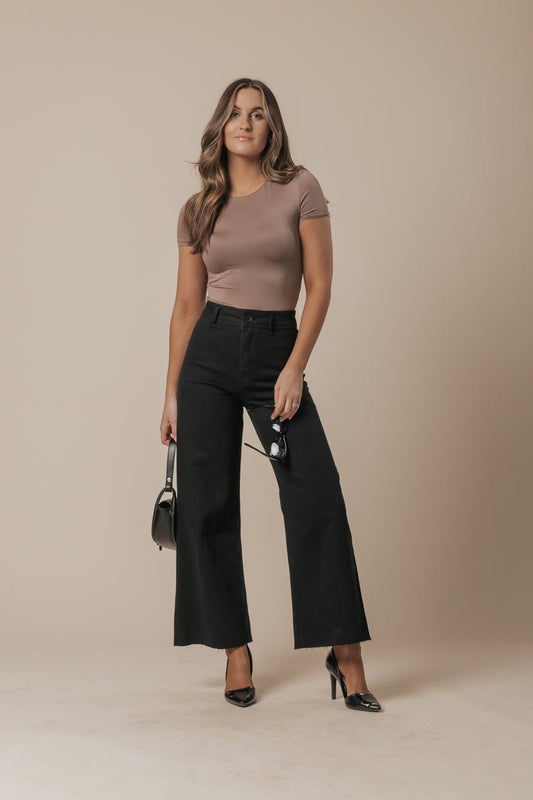 Woman in a taupe t-shirt, Black Straight Wide Leg Jeans, black heels, and bag poses against a beige background.