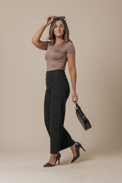 Woman in a taupe top and Black Straight Wide Leg Jeans holds a black bag and sunglasses, standing against a beige background.
