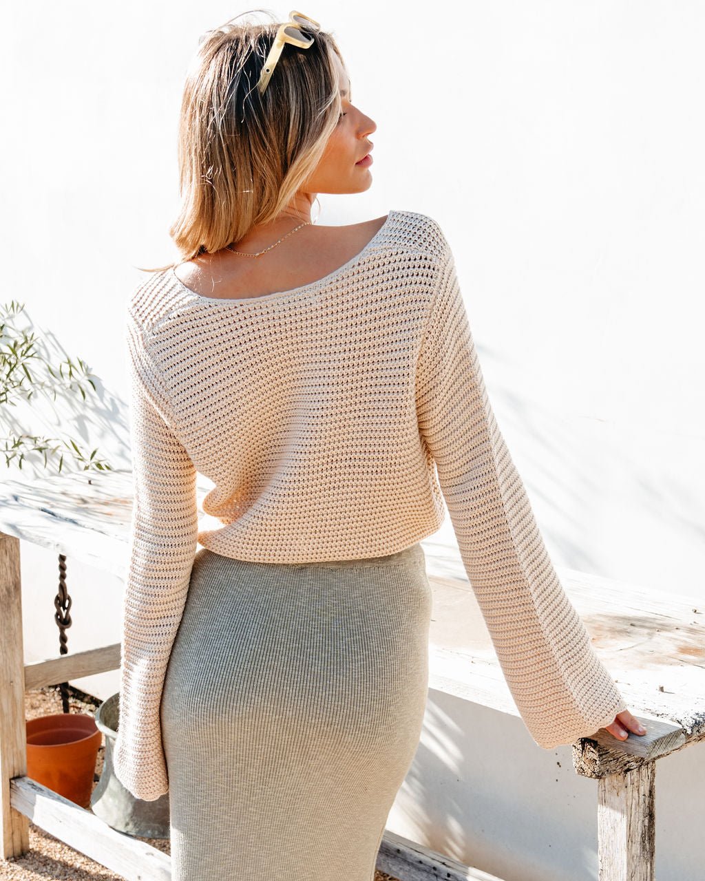 Woman in a California Cinched Button Down Sweater showcases relaxed vacation style by a rustic bench in bright sunlight.