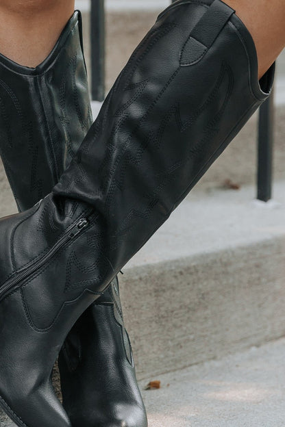 A person wearing Dirty Laundry Upwind Western Boots-FINAL SALE sits on concrete steps, showcasing their decorative stitching.