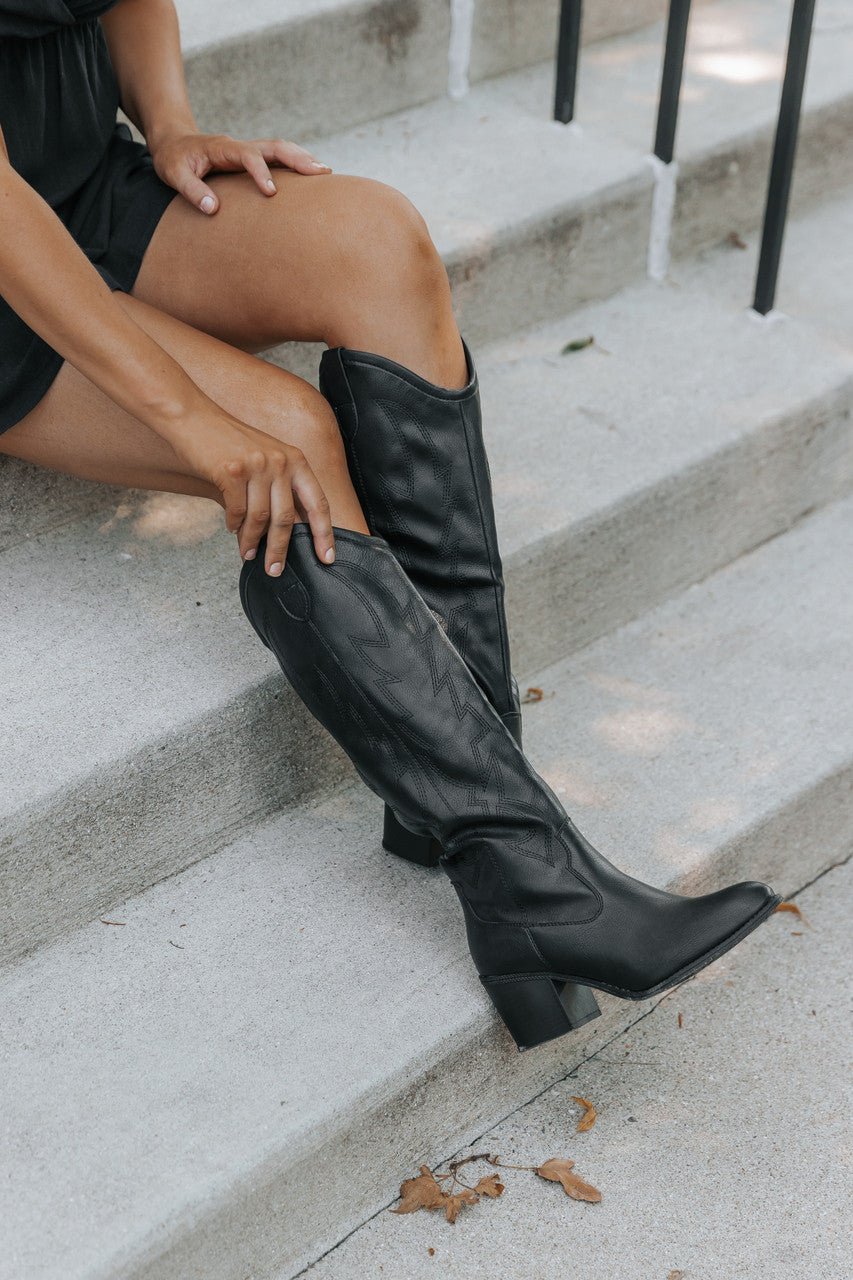 On concrete steps, a person in a black dress adjusts Dirty Laundry Upwind Western Boots with a bold block heel.