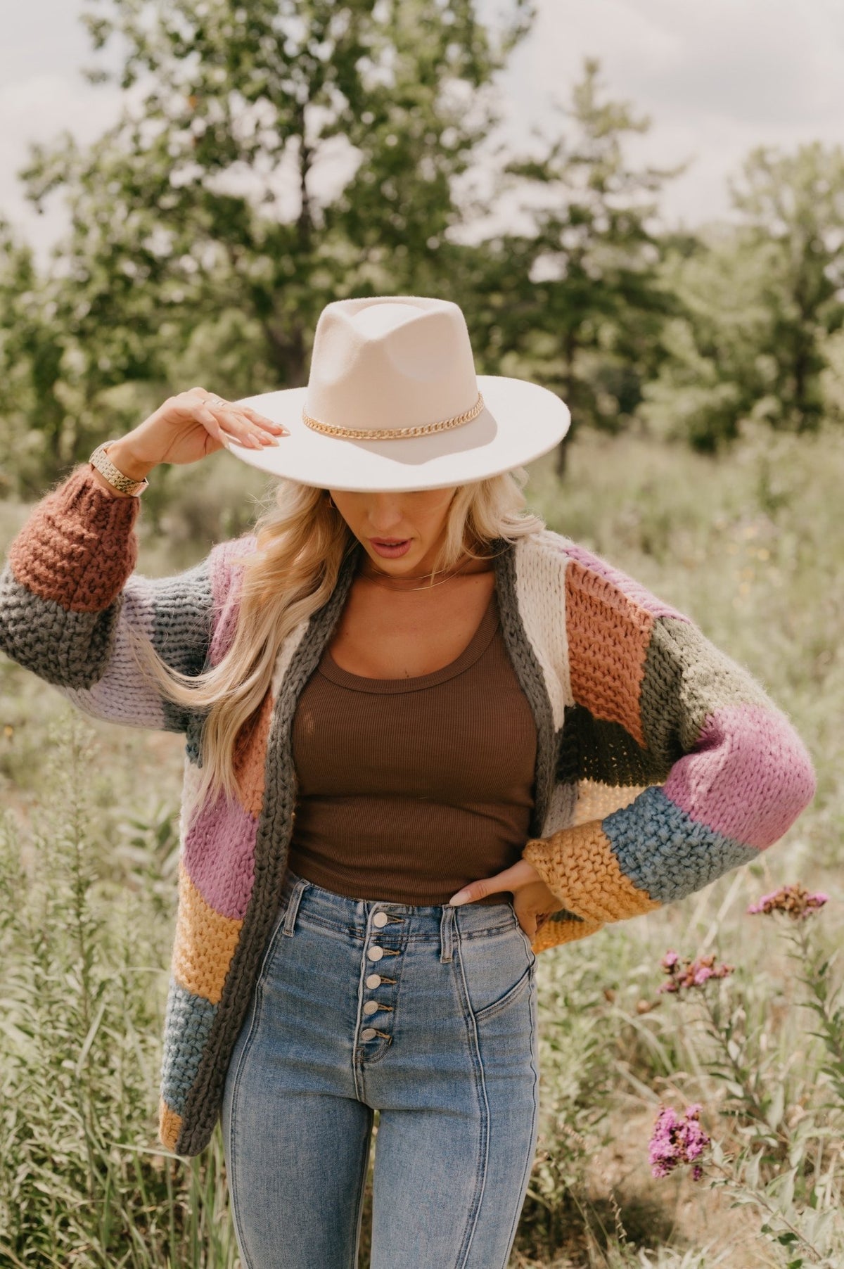 Woman in Falling Leaves Olive Color Block Cardigan, brown tank, and jeans stands outdoors holding her hat amid lush greenery.