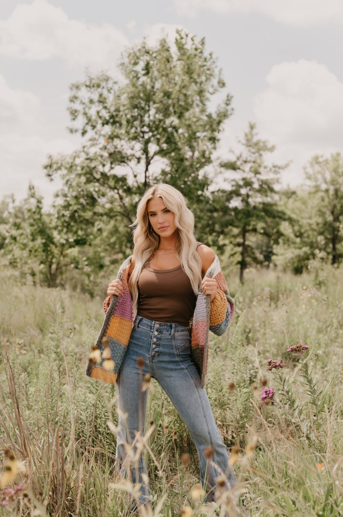 Woman with long blonde hair in jeans and a Falling Leaves Olive Color Block Cardigan stands in a wildflower-filled grassy field.