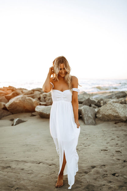 Barefoot woman in the Odella Off Shoulder Smocked Maxi Dress walks on a sandy beach at sunset, gently touching her hair.