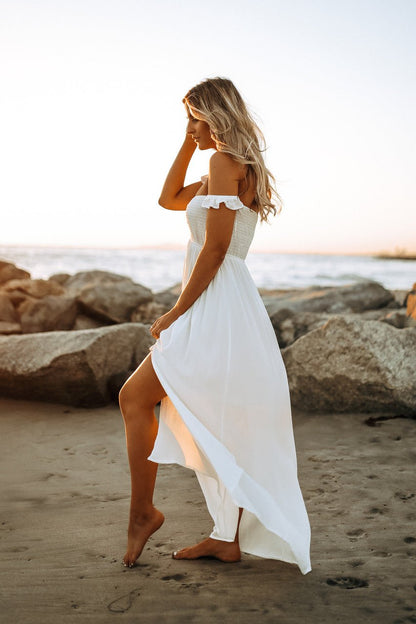 Barefoot woman in the Odella Off Shoulder Dress: Smocked Maxi in Off White walks on a sandy beach at sunset near rocks.