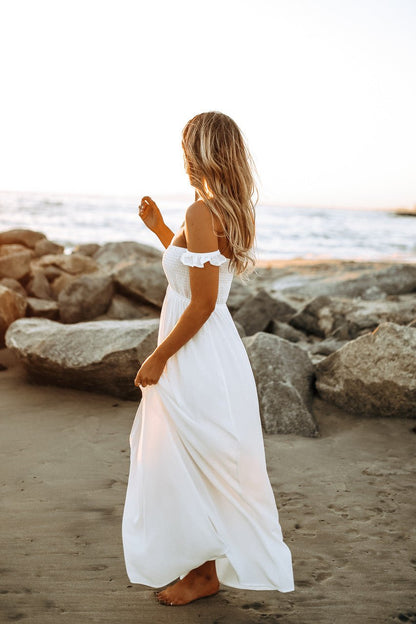 Barefoot woman in Odella Off Shoulder Dress stands on sandy beach at sunset, gazing at the ocean and rocks.