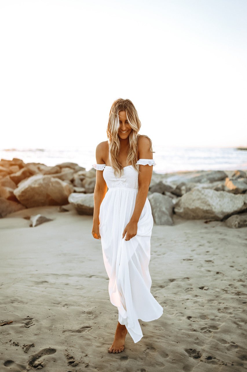 Woman in the Odella Off Shoulder Smocked Maxi Dress strolls barefoot on the beach at sunset, smiling with the ocean behind her.
