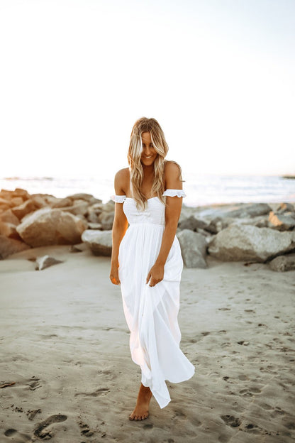 Woman in the Odella Off Shoulder Smocked Maxi Dress strolls barefoot on the beach at sunset, smiling with the ocean behind her.