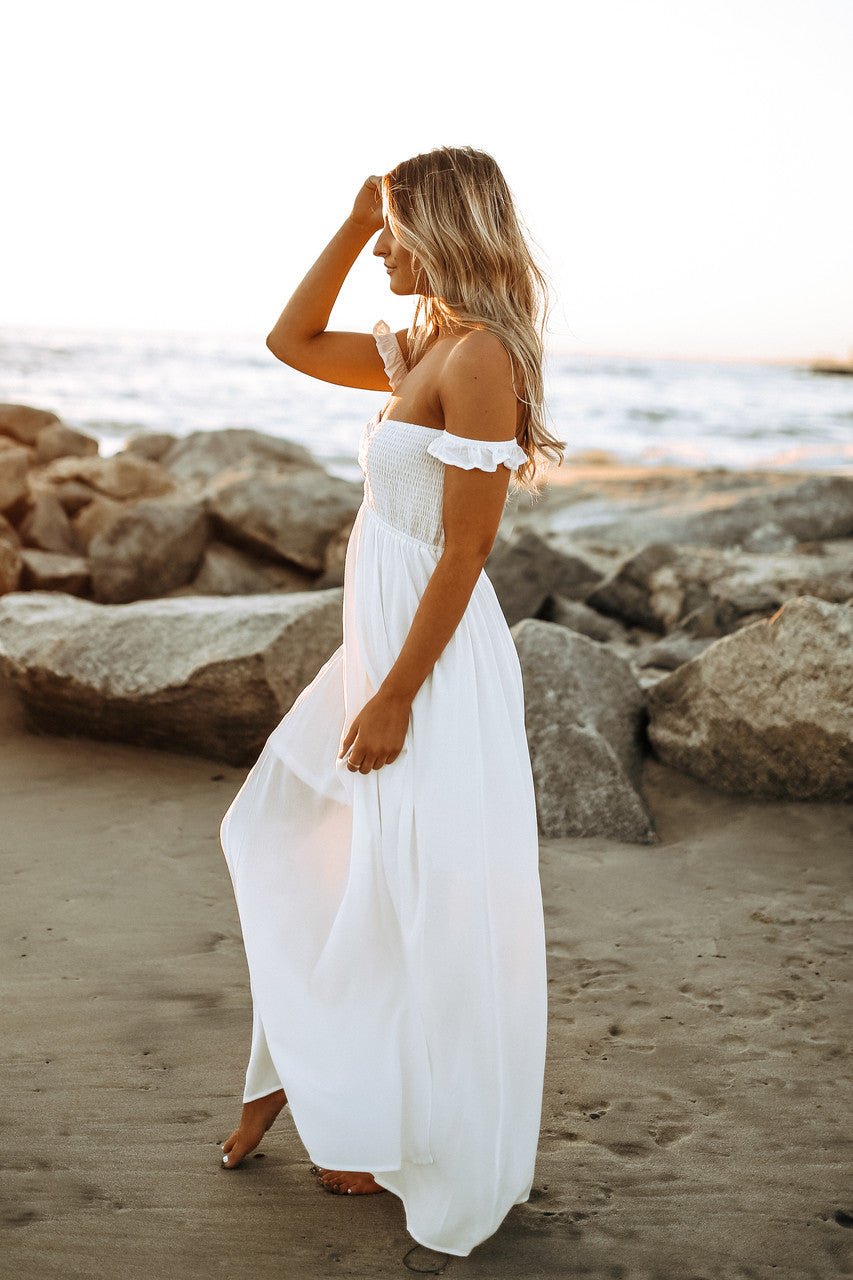 Barefoot woman in Odella Off Shoulder Smocked Maxi Dress stands on a beach at sunset, gazing toward the horizon near large rocks.