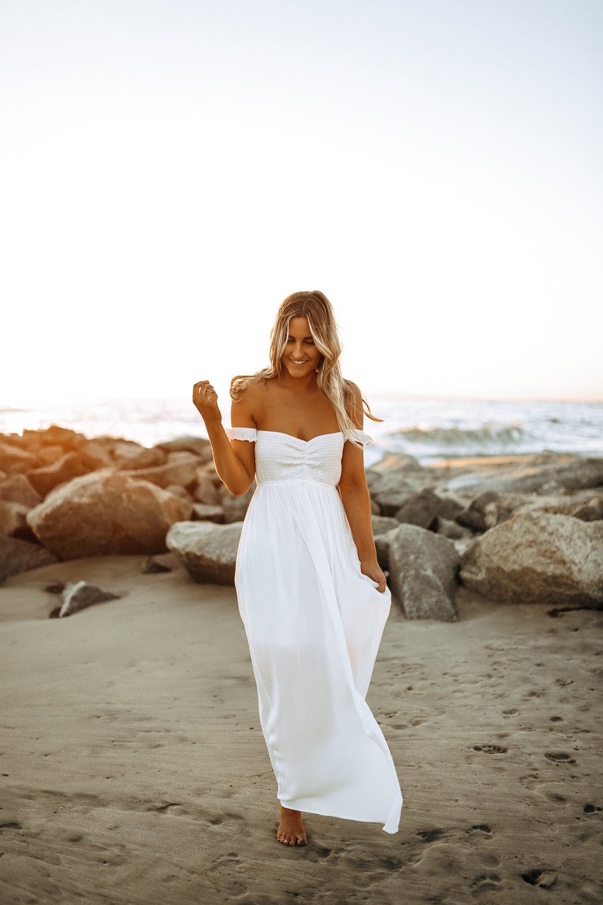 A woman in the Odella Off Shoulder Smocked Maxi Dress in off white walks barefoot on a sandy beach at sunset.