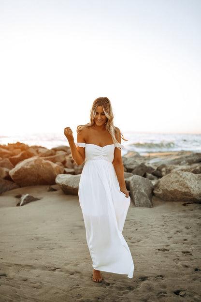 A woman in the Odella Off Shoulder Smocked Maxi Dress in off white walks barefoot on a sandy beach at sunset.