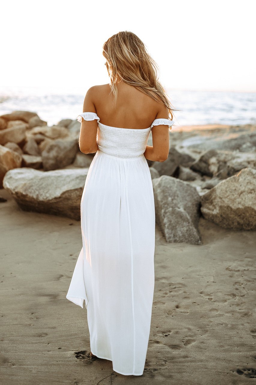 Woman in Odella Off Shoulder Dress: Smocked Maxi in Off White stands on a beach at sunset, gazing at the ocean near large rocks.