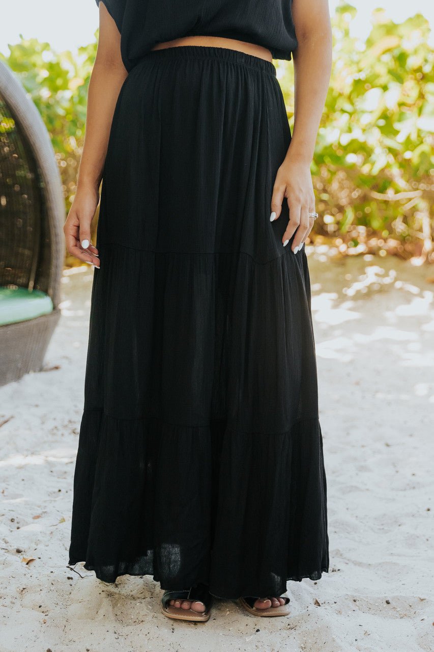 Person in the Sunrise Short Sleeve Top & Maxi Skirt Black Set, barefoot on sand near greenery.