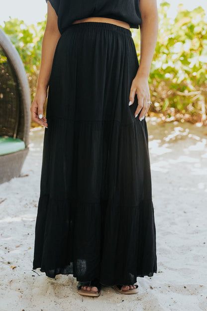 Person in the Sunrise Short Sleeve Top & Maxi Skirt Black Set, barefoot on sand near greenery.