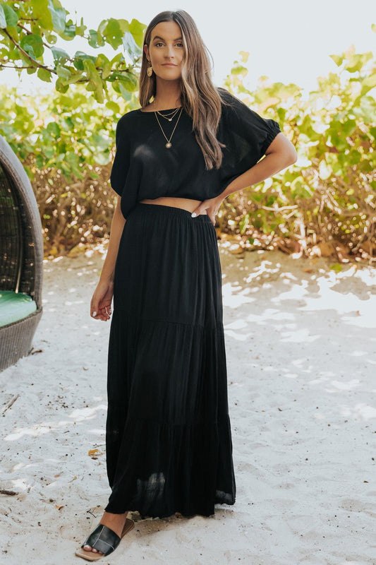 Woman models Sunrise Short Sleeve Top & Maxi Skirt Black Set-FINAL SALE, posing on sandy ground near greenery.