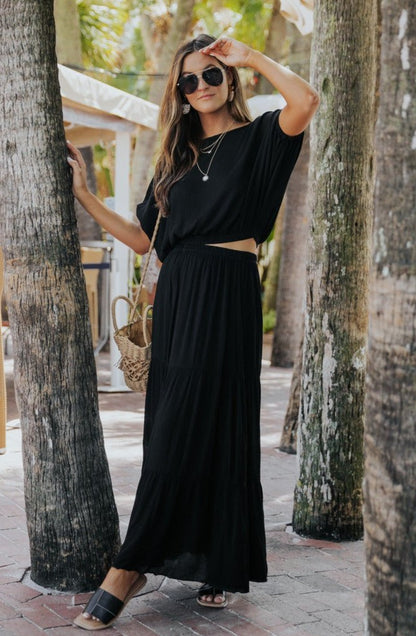Woman in the Sunrise Short Sleeve Top & Maxi Skirt Black Set stands by trees, wearing sunglasses and sandals, holding a straw bag.
