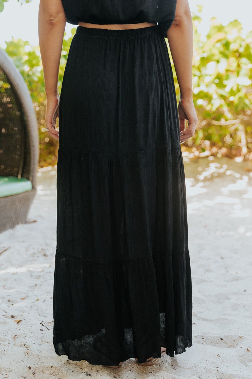 Person wearing the Sunrise Short Sleeve Top & Maxi Skirt Black Set-FINAL SALE, barefoot on sand with greenery in the background.