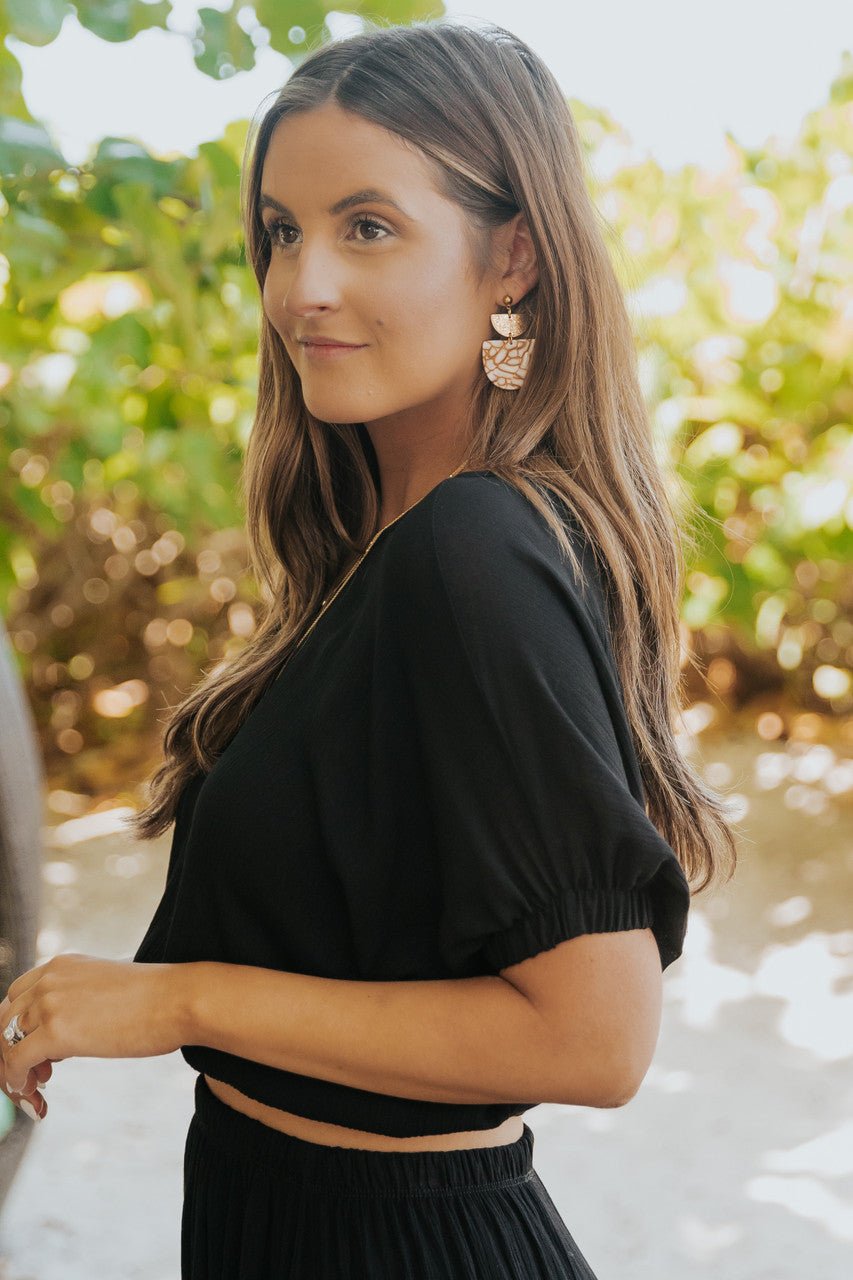 A woman in the Sunrise Short Sleeve Top & Maxi Skirt Black Set stands outdoors, looking to the side with greenery behind her.