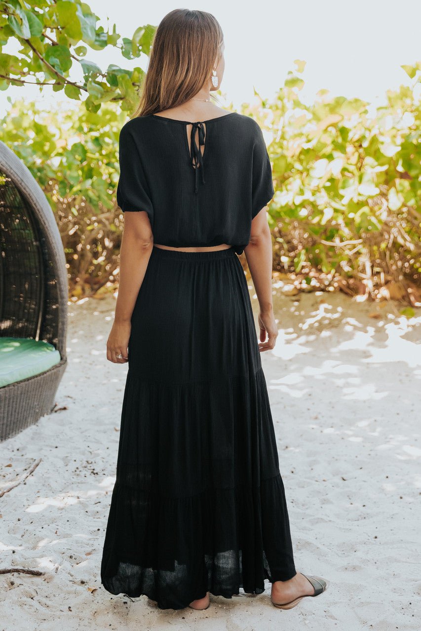 Woman wearing the Sunrise Short Sleeve Top & Maxi Skirt Black Set stands on sand, facing away, with green leaves and a wicker seat nearby.