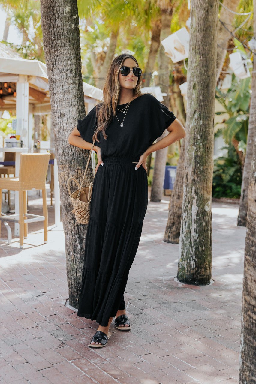 Woman in the Sunrise Short Sleeve Top & Maxi Skirt Black Set stands by palm trees with a straw bag, sunglasses, and sandals.