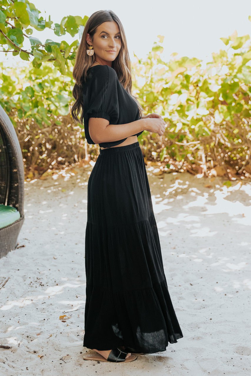 Woman wears the Sunrise Short Sleeve Top & Maxi Skirt Black Set, standing outdoors with greenery and sunlight behind her.