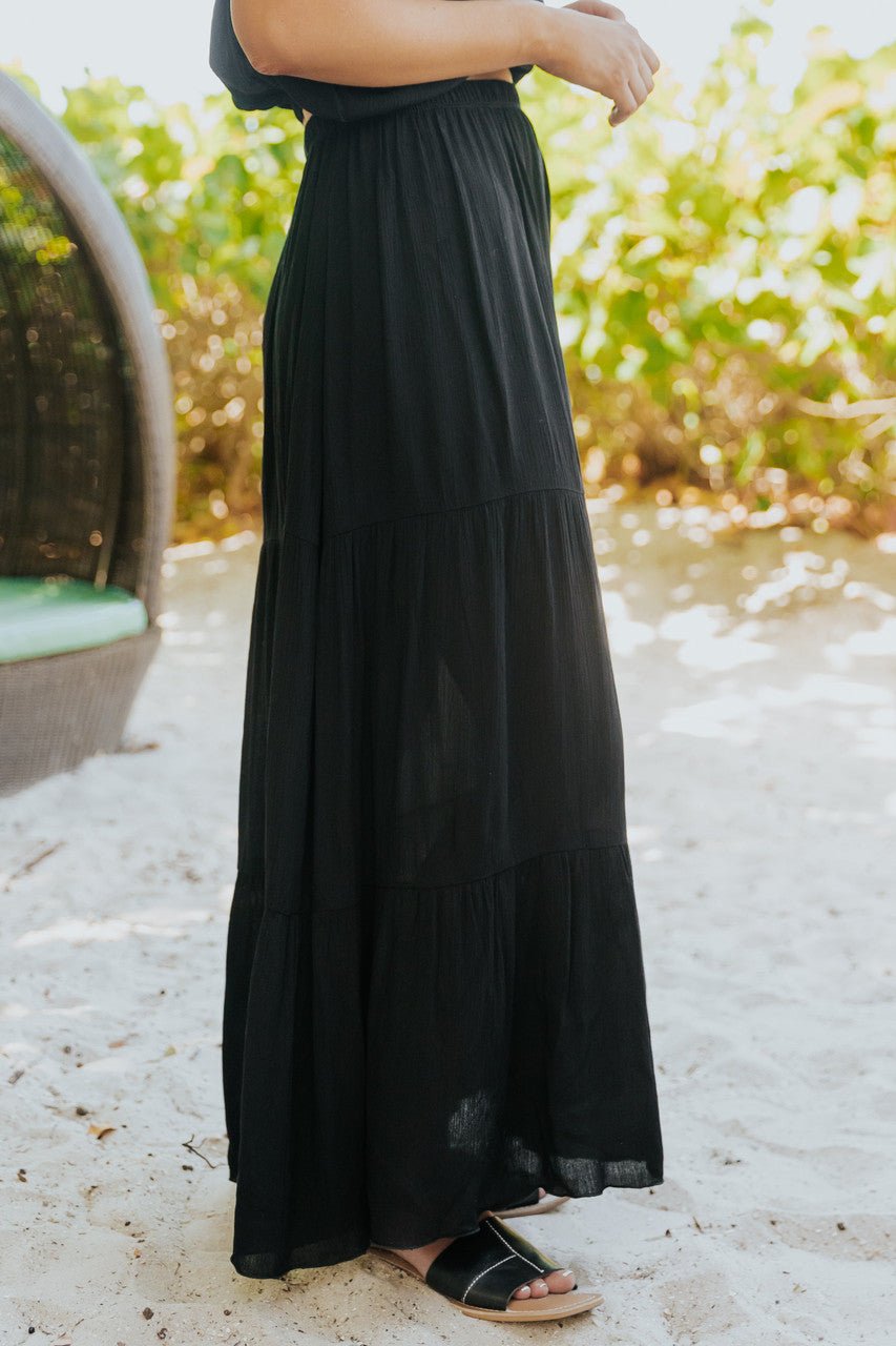 Woman wears the Sunrise Short Sleeve Top & Maxi Skirt Black Set on sandy ground, greenery in the background.