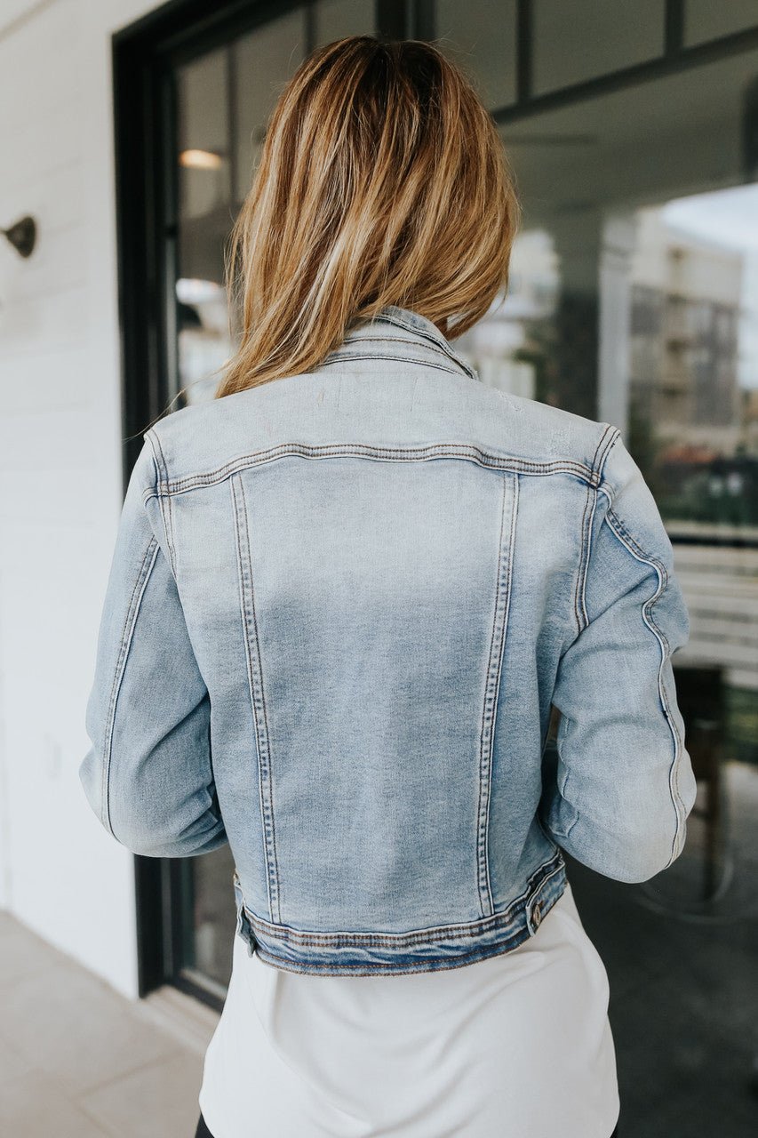 Someone with long hair wears the Sweet South Faded Denim Jacket - Light Wash, shown from behind indoors near glass doors for a casual look.
