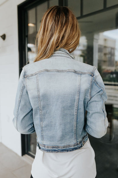 Someone with long hair wears the Sweet South Faded Denim Jacket - Light Wash, shown from behind indoors near glass doors for a casual look.