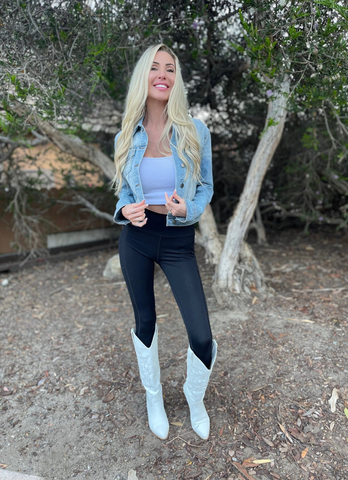 Woman with long blonde hair smiles outdoors by trees in the Sweet South Faded Denim Jacket - Light Wash and white boots.