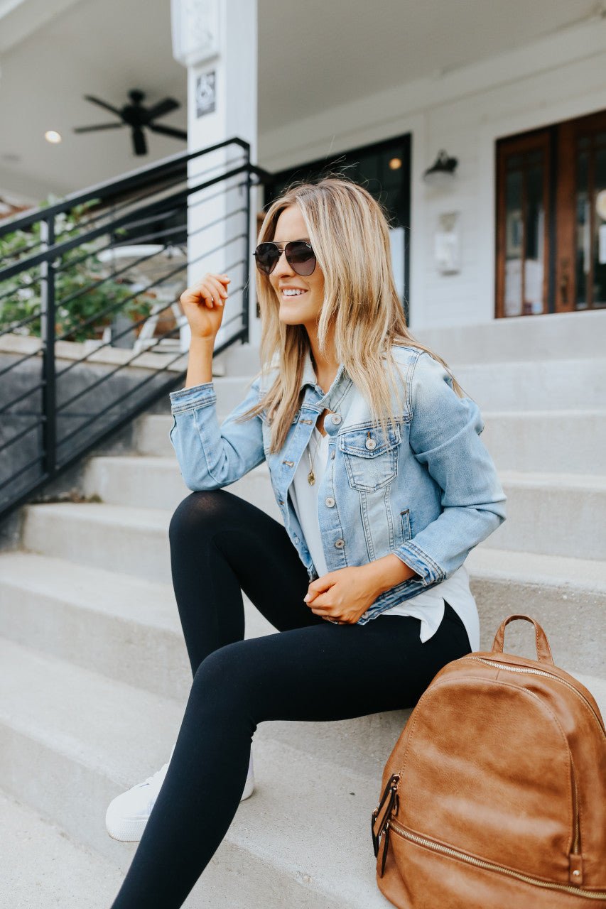 Woman in sunglasses and Sweet South Faded Denim Jacket—Light Wash sits on steps, smiling, brown backpack by her side.