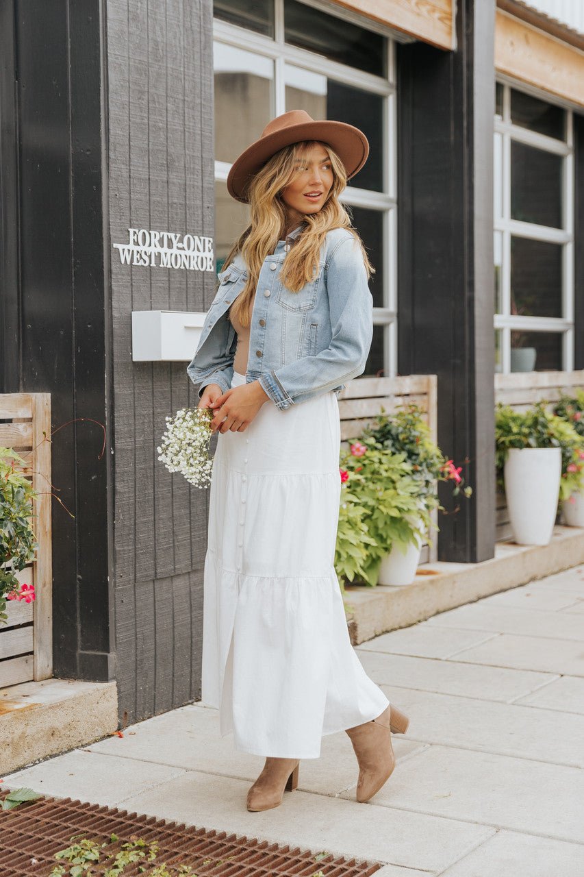 Woman in Sweet South Faded Denim Jacket - Light Wash layers over a white dress, holding small flowers beside potted plants.
