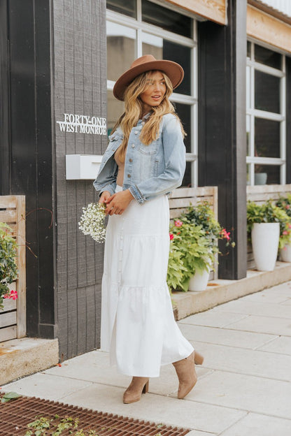 Woman in Sweet South Faded Denim Jacket - Light Wash layers over a white dress, holding small flowers beside potted plants.
