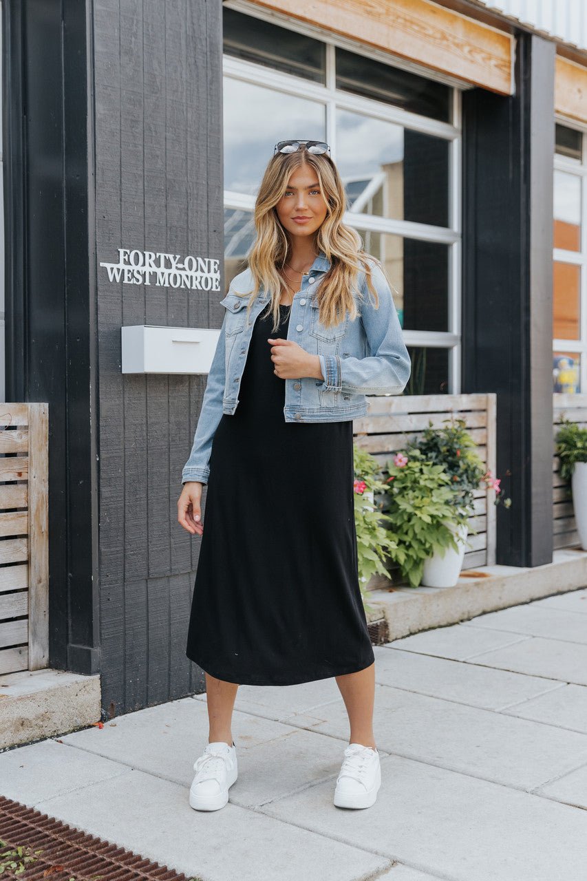 A woman stands outside a building in a black dress, Sweet South Faded Denim Jacket - Light Wash, white sneakers, and sunglasses.