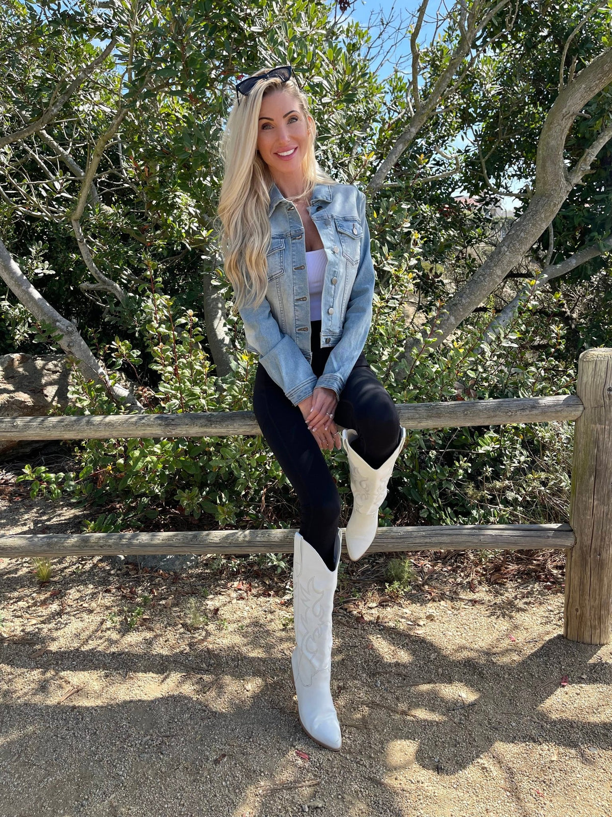 Woman with long blonde hair models Sweet South Faded Denim Jacket - Light Wash, black pants, and white boots by a wooden fence and green trees.