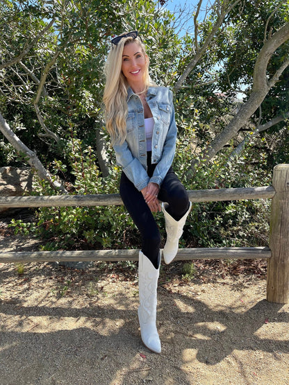 Woman with long blonde hair models Sweet South Faded Denim Jacket - Light Wash, black pants, and white boots by a wooden fence and green trees.